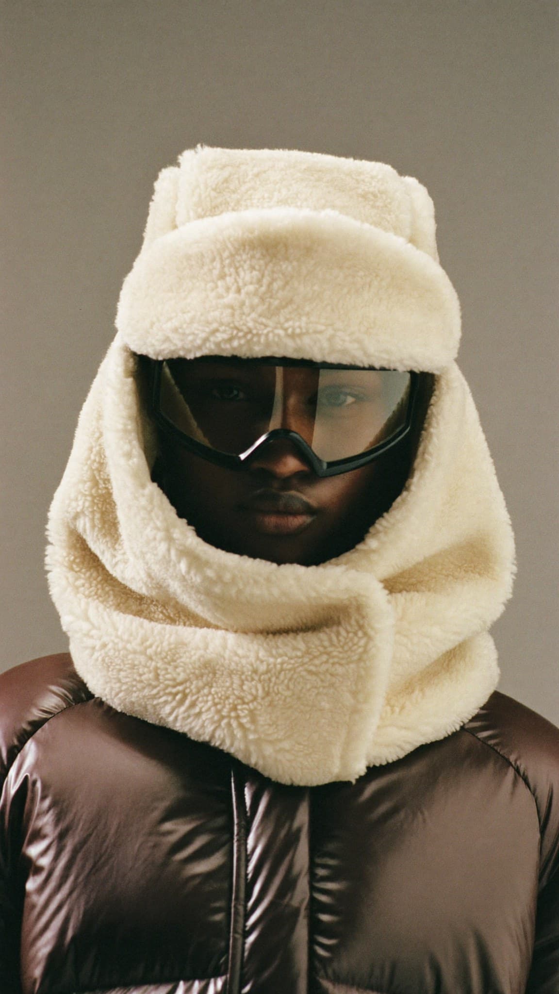The Shearling Hood: a deep cream shearling hood with a folded brim and a collar that wraps once, a clear visor set inside it, worn with a dark brown puffer jacket.
