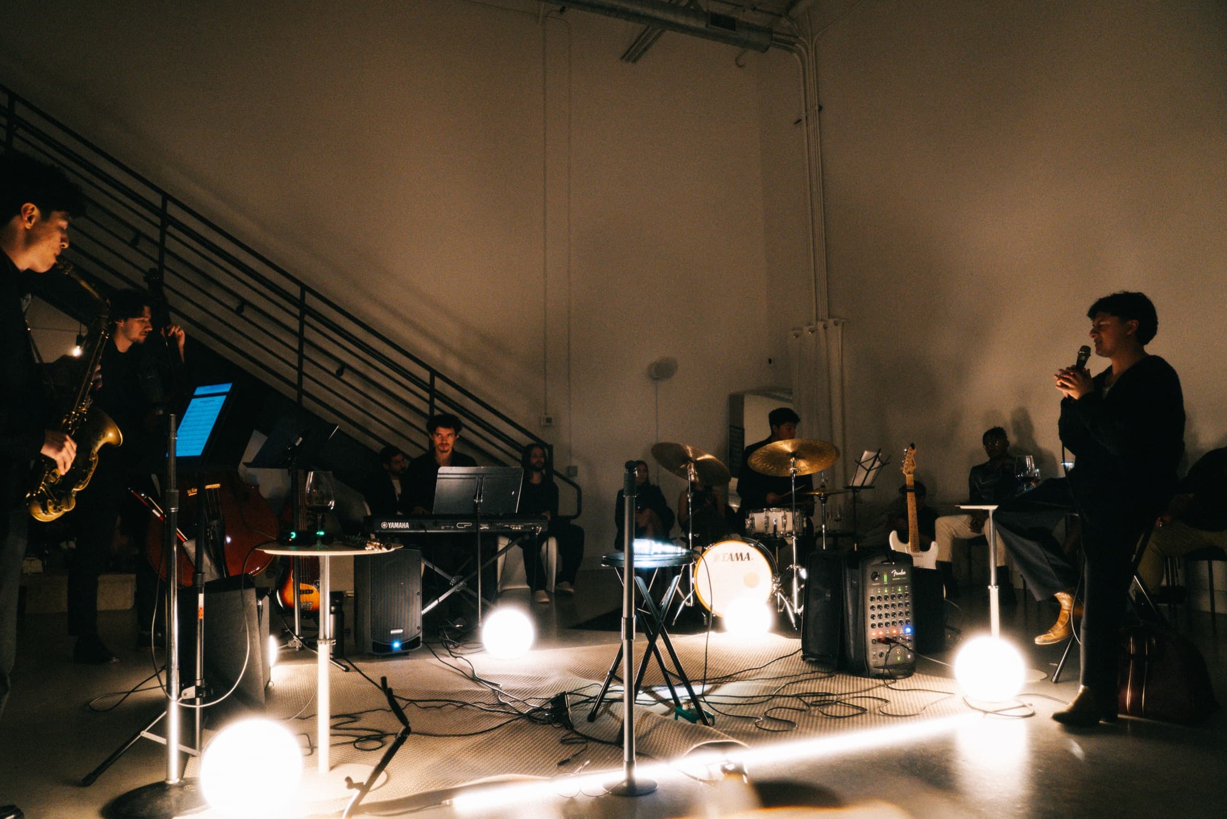 A five-piece band mid-set — saxophone and upright bass at left, vocalist at right, lit by floor globes.