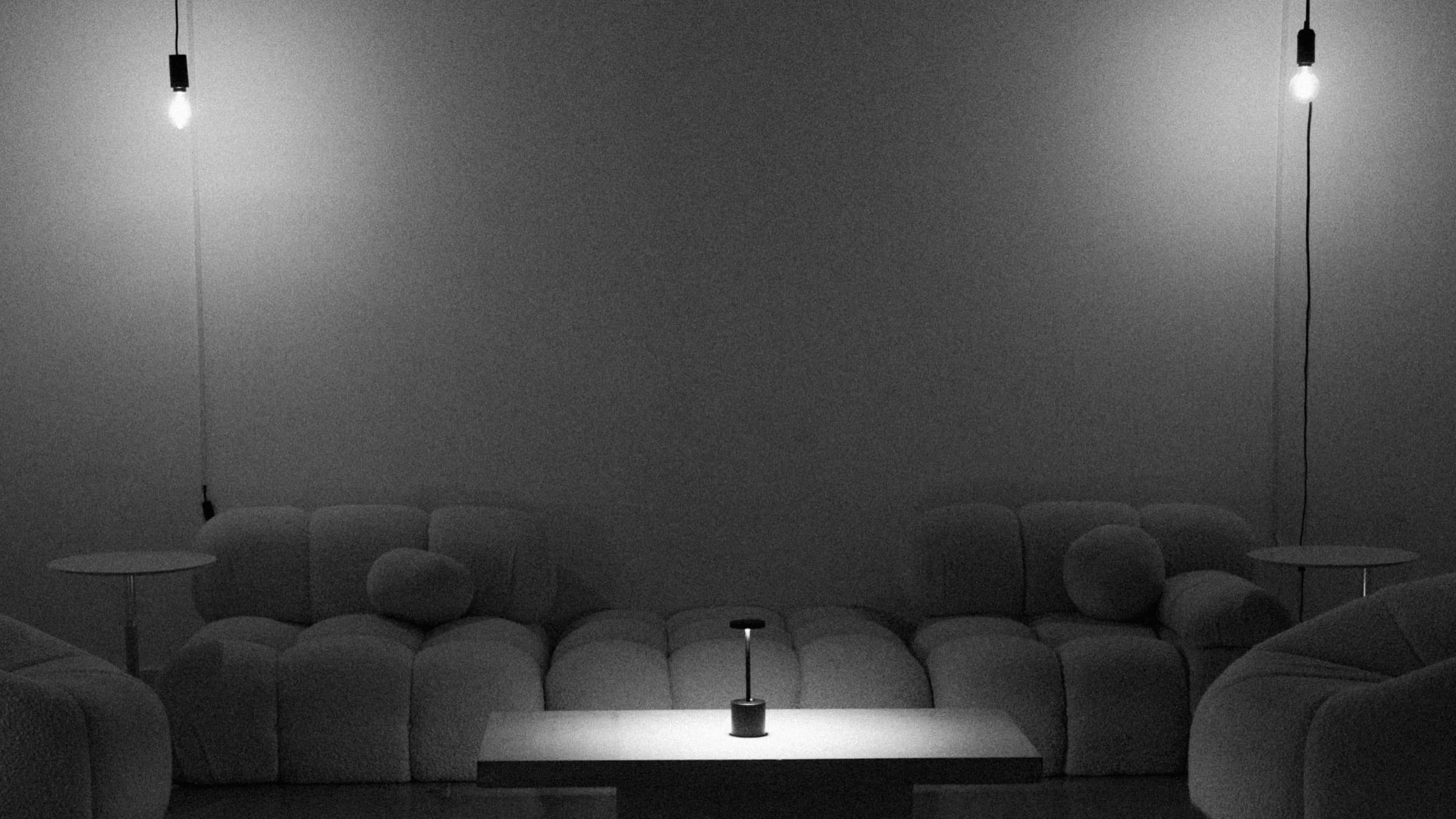 The lounge at night, empty — two bare pendant bulbs over low bouclé seating and a lit table lamp.
