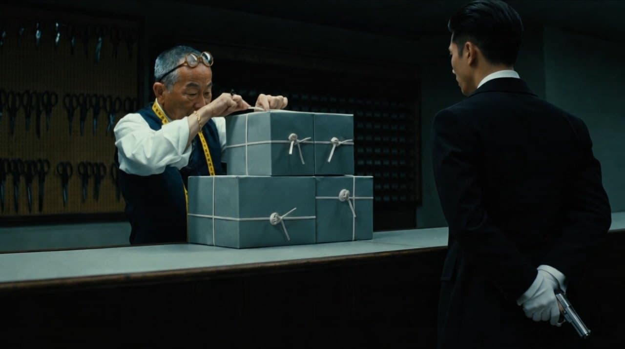 Scene 10: the old tailor tying grey boxes while a young man waits with a gloved hand behind his back.