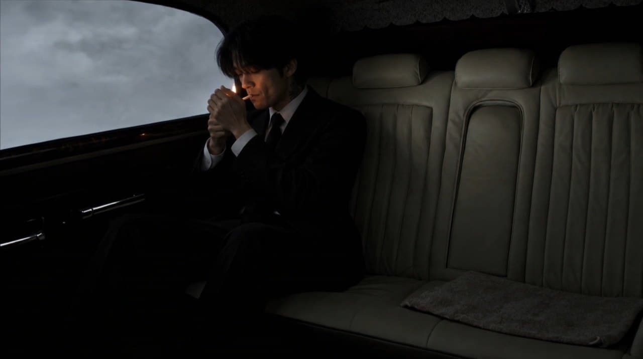 Scene 05: a young man alone on the long back seat, lighting a cigarette against the grey window.